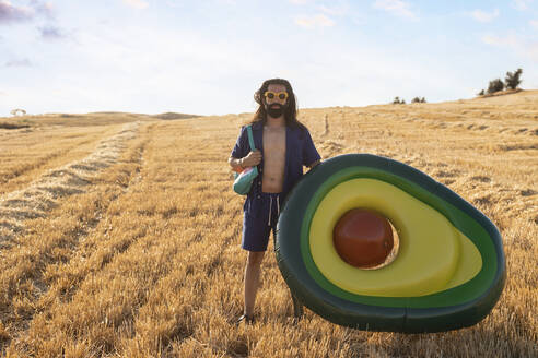 Bärtiger Mann mit Beinprothese und Sonnenbrille steht neben einem aufblasbaren Ring in Form einer Avocado auf einem Feld - JCCMF06964