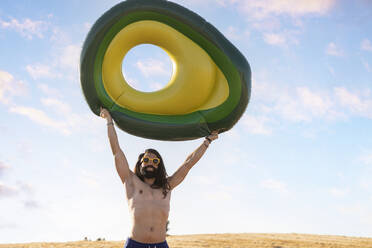 Lächelnder Hipster-Mann mit erhobener Hand, der einen aufblasbaren Ring an einem sonnigen Tag hält - JCCMF06966