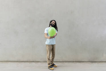 Smiling bearded man giving green balloon in front of wall - JCCMF06974