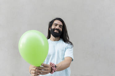 Happy hipster man with long black hair giving green balloon in front of wall - JCCMF06975