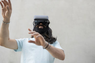 Lächelnder bärtiger Mann mit Virtual-Reality-Simulator vor einer Wand - JCCMF06986