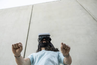 Smiling hipster man wearing virtual reality simulator gesturing fists in front of wall - JCCMF06988