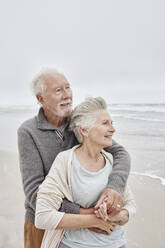 Happy senior couple standing smiling on windy beach - RORF03047