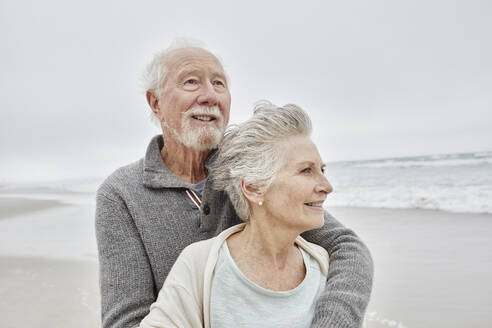 Happy senior couple standing smiling on windy beach - RORF03048