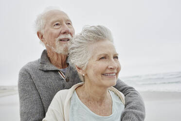 Happy senior couple standing smiling on windy beach - RORF03050