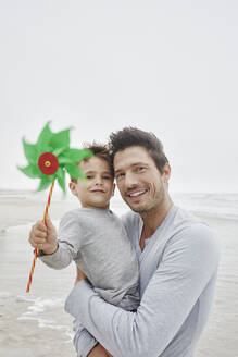 Vater trägt Sohn am Strand und hält grünes Windrädchen - RORF03053