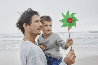 Vater trägt Sohn am Strand und hält grünes Windrädchen - RORF03055