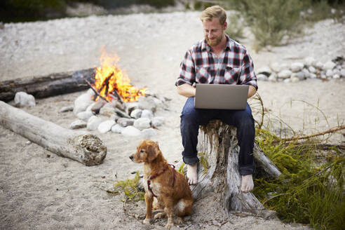 Lächelnder Geschäftsmann mit Laptop neben einem Hund vor einem Lagerfeuer - CVF02083