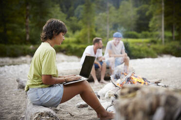 Freelancer using laptop sitting on fallen tree friends in background - CVF02084