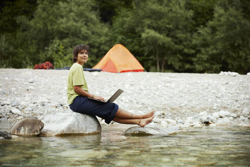 Lächelnde Geschäftsfrau mit Laptop auf einem Felsen am Flussufer sitzend - CVF02088