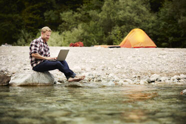 Lächelnder Geschäftsmann mit Laptop auf einem Felsen am Flussufer sitzend - CVF02089