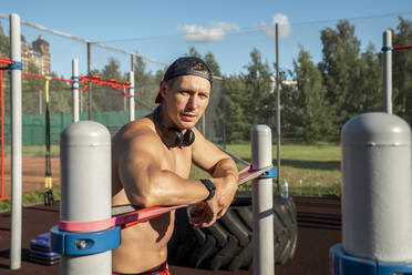 Ein fitter Mann mit Mütze steht mit einem Widerstandsband an einer horizontalen Stange im Park - VPIF06909