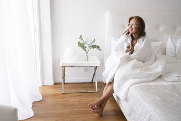 Lächelnde ältere Frau, eingewickelt in eine Decke und Tee trinkend, auf einem Bett sitzend - SVKF00429