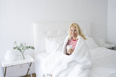 Smiling woman wrapped in blanket drinking tea in bed - SVKF00432