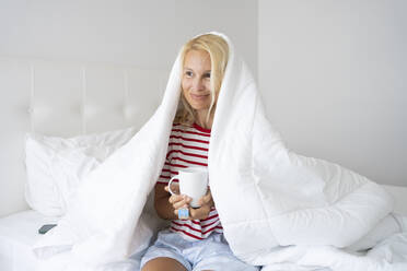 Smiling woman holding tea wrapped in blanket on bed - SVKF00433