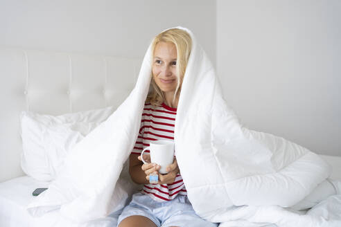 Lächelnde Frau mit Tee in einer Decke eingewickelt auf einem Bett - SVKF00433