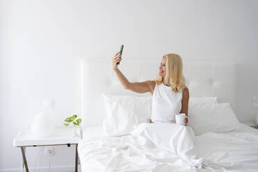Frau nimmt Selfie durch Handy sitzend auf Bett - SVKF00461