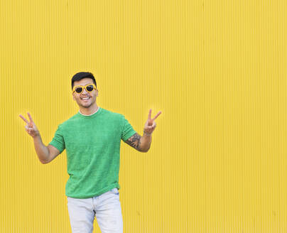 Happy young man gesturing peace sign in front of yellow wall - JCCMF07003
