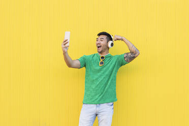 Happy young man showing biceps on video call through smart phone in front of yellow wall - JCCMF07004