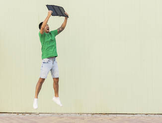 Happy young man jumping with solar panel in front of wall - JCCMF07010