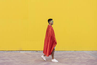 Young man wearing red superhero cape walking in front of yellow wall - JCCMF07014