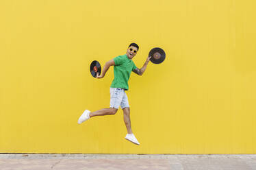 Young man jumping with vinyl records in front of wall - JCCMF07022