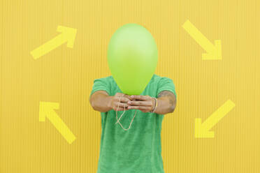 Young man covering face with green balloon in front of arrow symbols on yellow wall - JCCMF07026