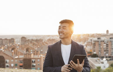 Happy young businessman with tablet PC standing in front of city - JCCMF07036