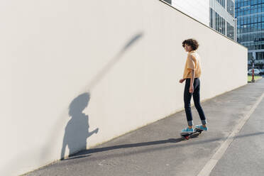 Frau, die an einem sonnigen Tag auf dem Fußweg an einer Mauer Skateboard fährt - MEUF07574