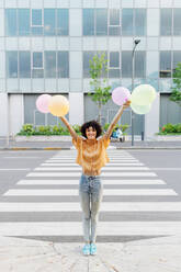 Lächelnde Frau mit erhobenen Armen, die Luftballons am Zebrastreifen auf der Straße hält - MEUF07638