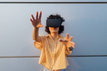 Junge Frau mit Virtual-Reality-Headset gestikuliert vor einer grauen Wand - MEUF07643