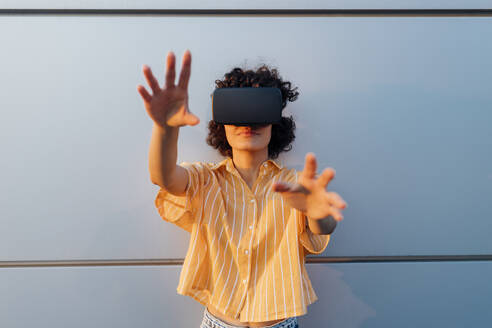 Junge Frau mit Virtual-Reality-Headset gestikuliert vor einer grauen Wand - MEUF07643