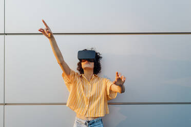 Young woman wearing virtual reality headset gesturing in front of wall - MEUF07644