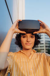 Young woman holding virtual reality headset by wall - MEUF07646