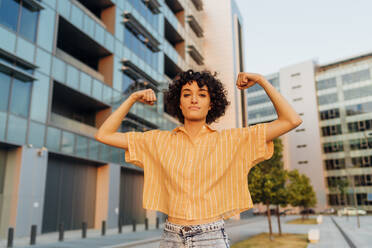 Confident young woman flexing muscles in front of buildings - MEUF07654