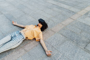 Carefree woman with arms outstretched wearing virtual reality headset lying on footpath - MEUF07666