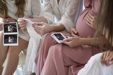 Hands of pregnant woman with friends holding ultrasound scan result at baby shower - ABIF01802