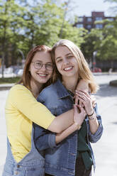 Smiling young woman embracing friend from behind on footpath - KMKF01880