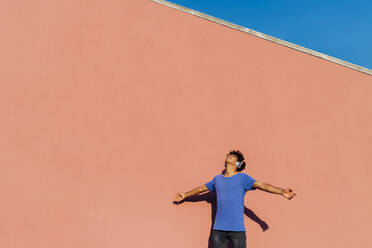 Man wearing headphones standing with arms outstretched on sunny day - MEUF07692