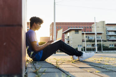 Junger Mann studiert mit Laptop auf dem Fußweg in der Stadt bei Sonnenuntergang - MEUF07741