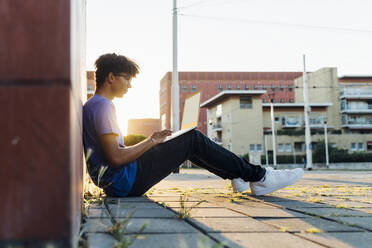 Young man studying through laptop sitting on footpath in city at sunset - MEUF07741