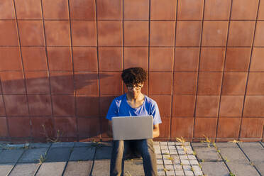 Junger Mann mit Laptop zum Lernen sitzend an die Wand gelehnt - MEUF07744
