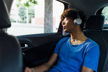 Junger Mann hört Musik über drahtlose Kopfhörer und schaut aus dem Autofenster - MEUF07746