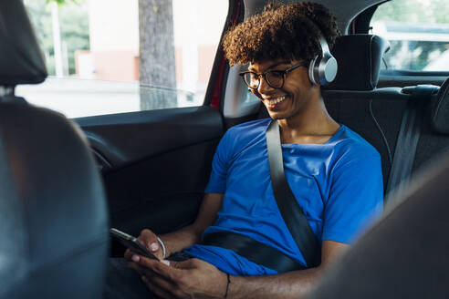 Lächelnder junger Mann mit drahtlosen Kopfhörern, der eine SMS über sein Smartphone schreibt - MEUF07747