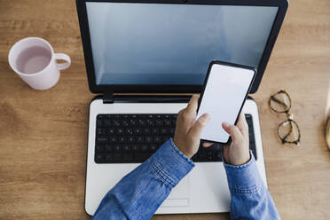 Hände einer Geschäftsfrau, die ein Smartphone über einem Laptop am Schreibtisch benutzt - EBBF05741