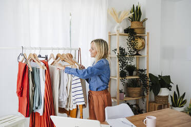 Lächelnde Modedesignerin hängt Kleidung auf einem Gestell im Büro auf - EBBF05747