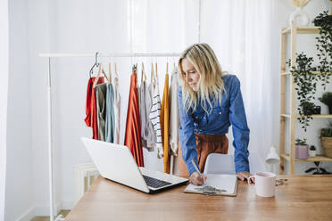 Modedesignerin schreibt auf einem Klemmbrett und schaut auf einen Laptop im Büro zu Hause - EBBF05750