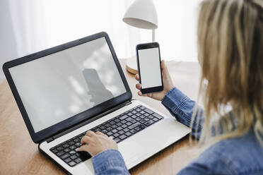 Geschäftsfrau arbeitet an einem Laptop und hält ein Smartphone im Büro zu Hause - EBBF05764