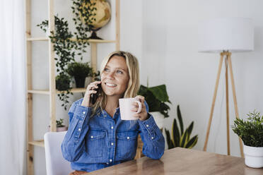 Nachdenkliche Geschäftsfrau, die mit ihrem Smartphone telefoniert und eine Tasse im Büro zu Hause hält - EBBF05768