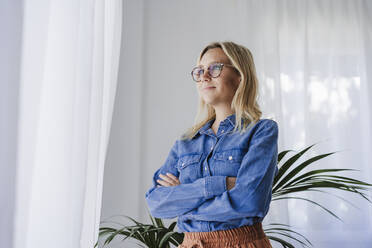 Young woman with arms crossed standing by window at home - EBBF05771